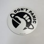 Circular black‑and‑white emblem featuring “DON’T PANIC” text and a stylised hand giving a thumbs‑up over a ringed planet, designed by Boon Hill Designs.
